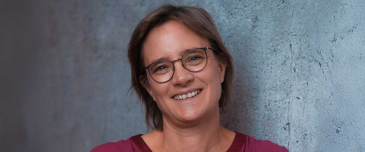 Carolin Pauly kurzes hellbraunes Haar, Brille, steht lächelnd vor Wand